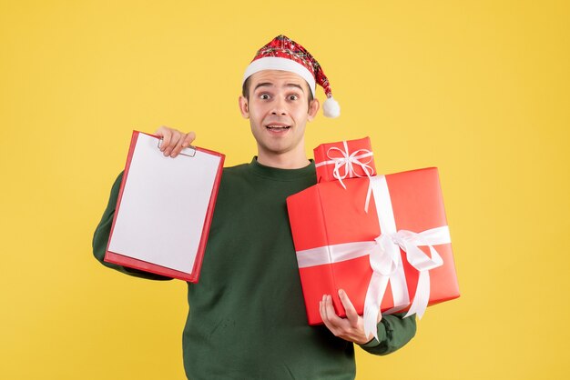 Vista frontal joven con gorro de Papá Noel con regalo y portapapeles de pie sobre fondo amarillo