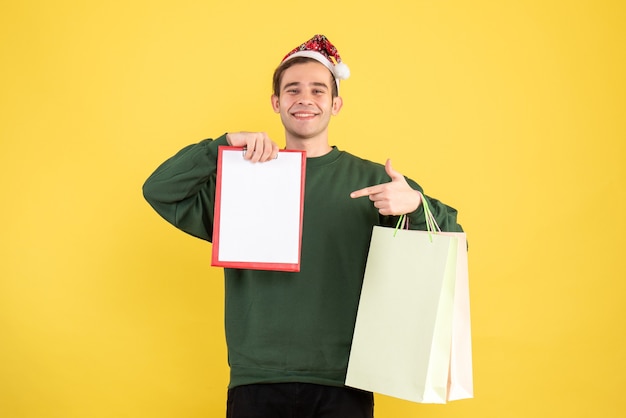 Vista frontal joven con gorro de Papá Noel sosteniendo bolsas de compras y portapapeles de pie sobre fondo amarillo espacio de copia