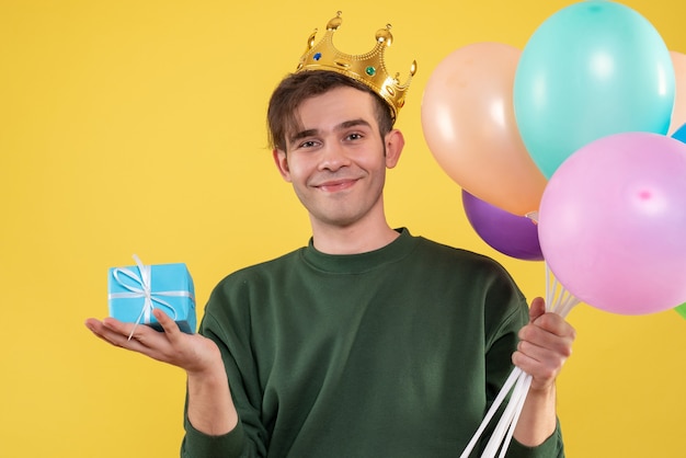 Vista frontal joven guapo con corona sosteniendo globos y caja de regalo azul sobre amarillo