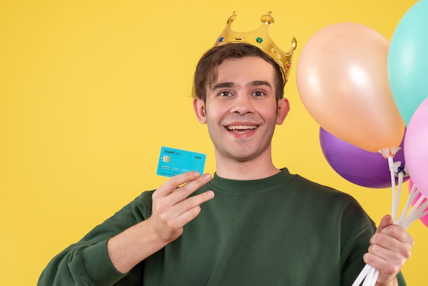 Vista frontal joven guapo con corona sosteniendo globos y tarjeta en amarillo