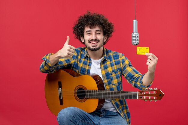 Vista frontal joven con guitarra sosteniendo una tarjeta bancaria en la pared roja cantante de banda de música en vivo color dinero