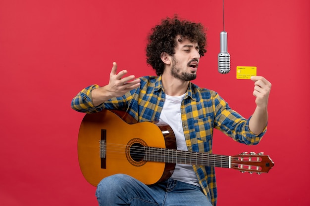 Vista frontal joven con guitarra sosteniendo una tarjeta bancaria en la pared roja en vivo músico concierto banda dinero color