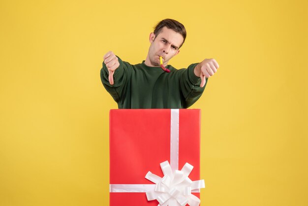 Vista frontal joven haciendo signo de pulgar hacia abajo de pie detrás de caja de regalo grande en amarillo