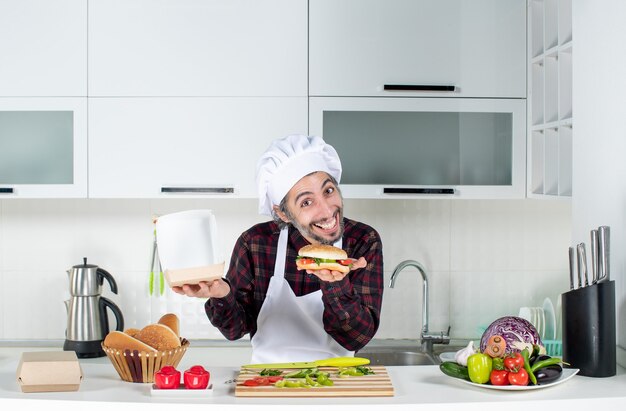 Vista frontal del joven hambriento sosteniendo una hamburguesa de pie detrás de la mesa de la cocina