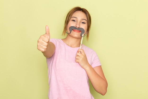 Una vista frontal joven hermosa en jeans azul camiseta rosa posando con bigote falso y sonriendo en verde