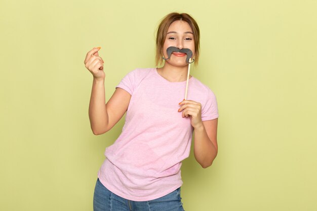 Una vista frontal joven hermosa en jeans azul camiseta rosa posando con bigote falso en verde