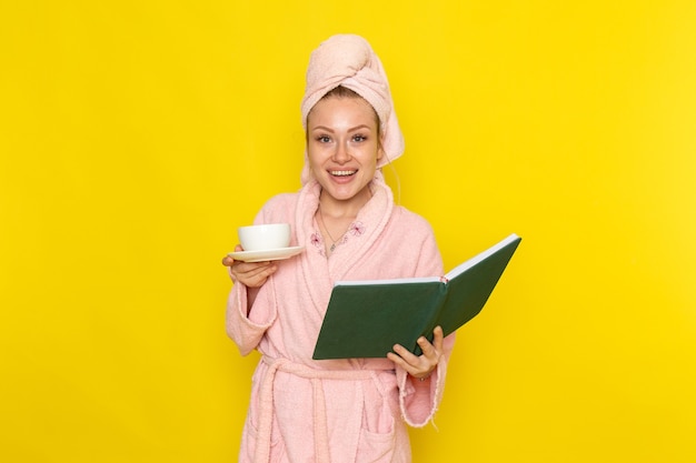 Vista frontal joven hermosa mujer en bata de baño rosa sosteniendo una taza de té y cuaderno verde