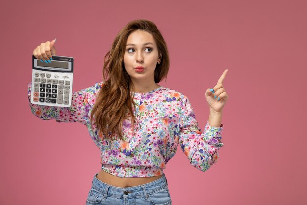 Vista frontal joven hermosa mujer en camisa diseñada con flores y jeans azul con calculadora en el fondo rosa