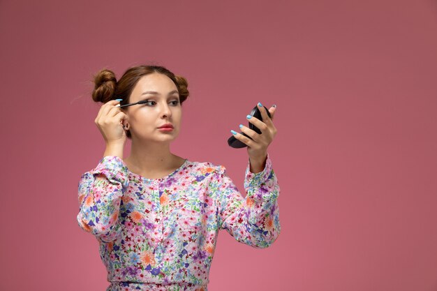 Vista frontal joven hermosa mujer en camisa de flor diseñada y jeans azul haciendo maquillaje en el fondo rosa