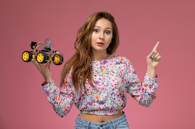 Vista frontal joven hermosa mujer en camisa de flor diseñada y jeans con coche de juguete posando sobre fondo rosa