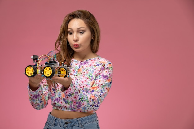 Vista frontal joven hermosa mujer en camisa de flor diseñada y jeans con coche de juguete posando sobre fondo rosa