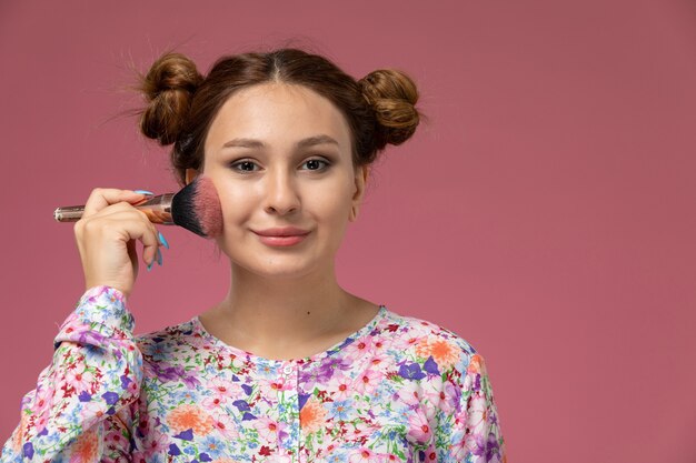 Vista frontal joven hermosa mujer en camisa de flor diseñada y jeans haciendo un maquillaje con una leve sonrisa sobre fondo rosa