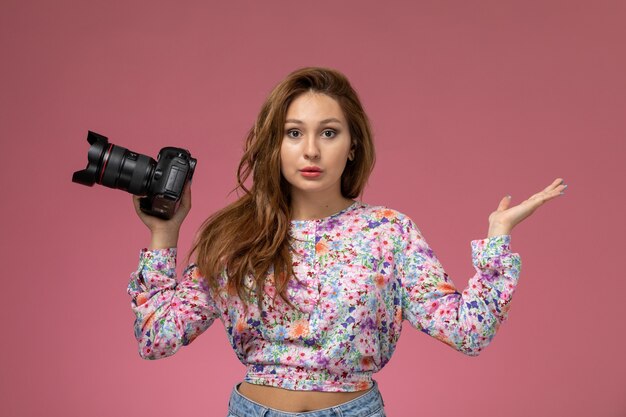 Vista frontal joven hermosa mujer en camisa de flor diseñada y jeans sosteniendo cámara de fotos sobre fondo rosa