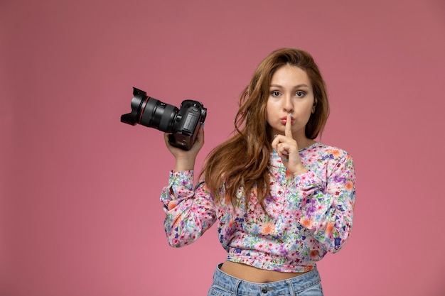 Vista frontal joven hermosa mujer en camisa de flor diseñada y jeans sosteniendo cámara de fotos sobre fondo rosa