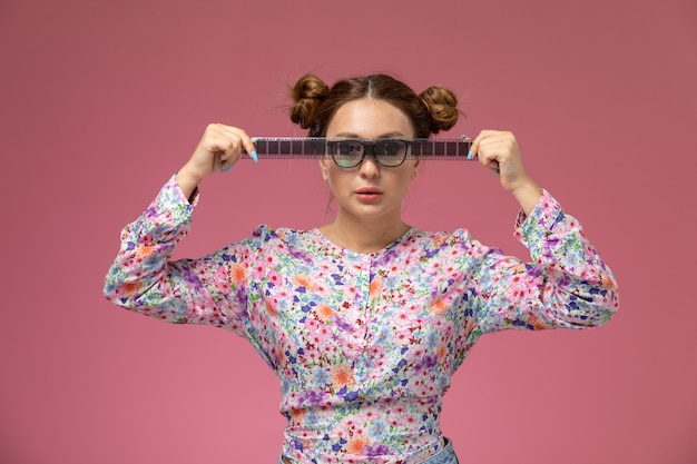 Foto gratuita vista frontal joven hermosa mujer en camisa de flor diseñada y jeans sosteniendo cinta de cine vieja en el fondo rosa