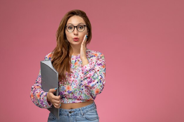 Vista frontal joven hermosa mujer en camisa de flor diseñada y jeans sosteniendo documento gris sobre fondo rosa