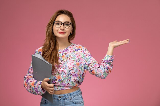 Vista frontal joven hermosa mujer en camisa de flor diseñada y jeans sosteniendo documento sobre fondo rosa