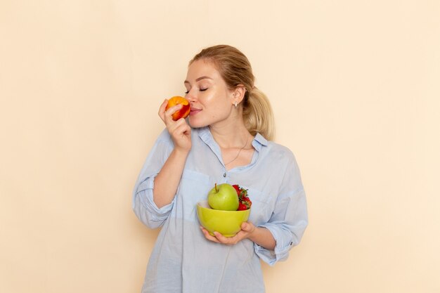 Vista frontal joven hermosa mujer en camisa sosteniendo placa con frutas que huelen a manzana en la pared de crema fruta madura modelo mujer pose