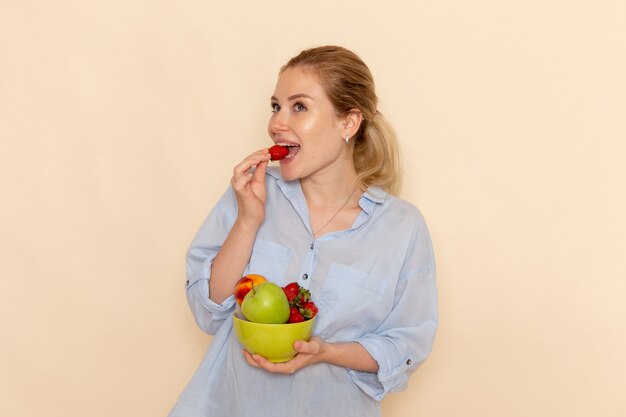 Vista frontal joven hermosa mujer en camisa sosteniendo plato con frutas comiendo fresa en la pared de crema fruta madura modelo mujer pose