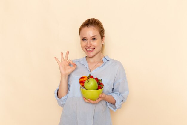 Vista frontal joven hermosa mujer en camisa sosteniendo un plato con frutas mostrando bien signo en la pared crema fruta madura modelo mujer pose