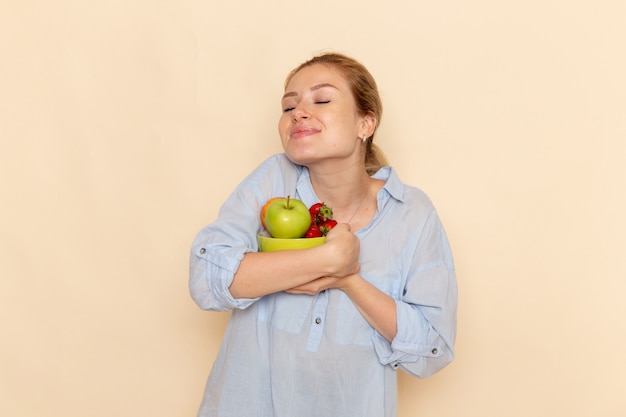 Vista frontal joven hermosa mujer en camisa sosteniendo el plato con frutas en la pared de crema ligera modelo de fruta pose de mujer
