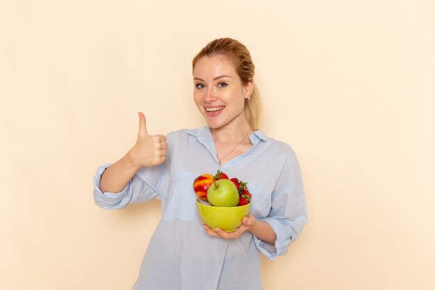 Vista frontal joven hermosa mujer en camisa sosteniendo plato con frutas con sonrisa en la pared crema fruta madura modelo mujer pose