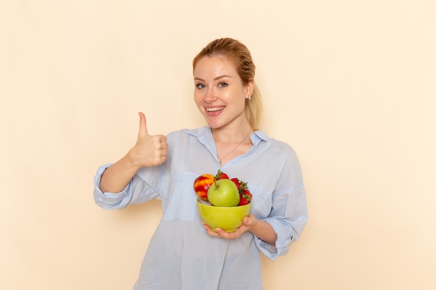 Vista frontal joven hermosa mujer en camisa sosteniendo plato con frutas con sonrisa en la pared crema fruta madura modelo mujer pose