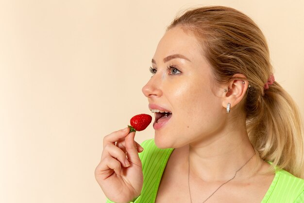 Vista frontal joven hermosa mujer en camisa verde comiendo fresas en la pared crema ligera modelo de fruta pose de mujer