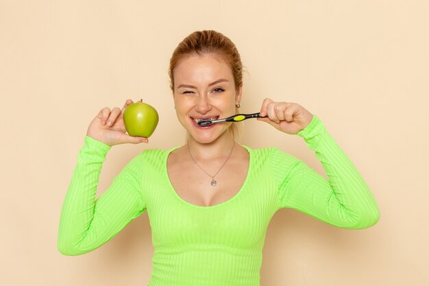 Vista frontal joven hermosa mujer en camisa verde con manzana verde limpiando sus dientes en la pared de crema modelo de fruta mujer suave
