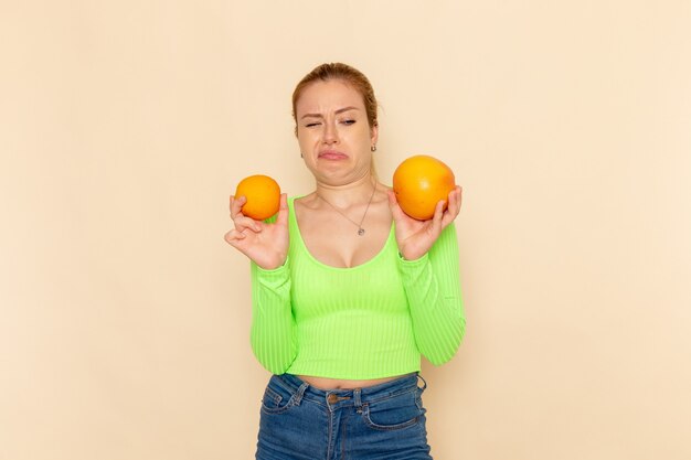 Vista frontal joven hermosa mujer en camisa verde con naranjas suaves frescas de diferentes tamaños en la pared crema modelo de fruta mujer suave