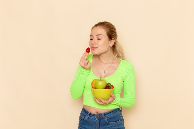 Vista frontal joven hermosa mujer en camisa verde con plato con frutas comiendo fresas en la pared de crema ligera modelo de fruta pose de mujer
