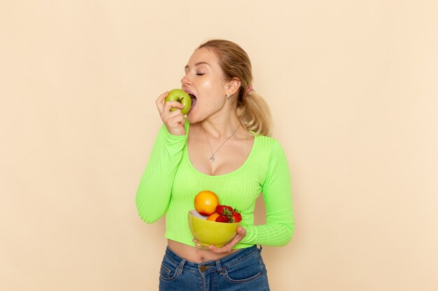 Vista frontal joven hermosa mujer en camisa verde con plato con frutas comiendo manzana en la pared de crema ligera modelo de fruta pose de mujer