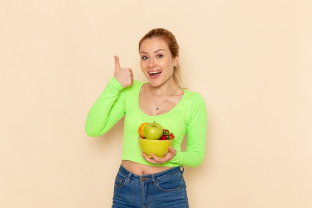 Vista frontal joven hermosa mujer en camisa verde con plato con frutas en la pared de crema ligera modelo de fruta pose de mujer