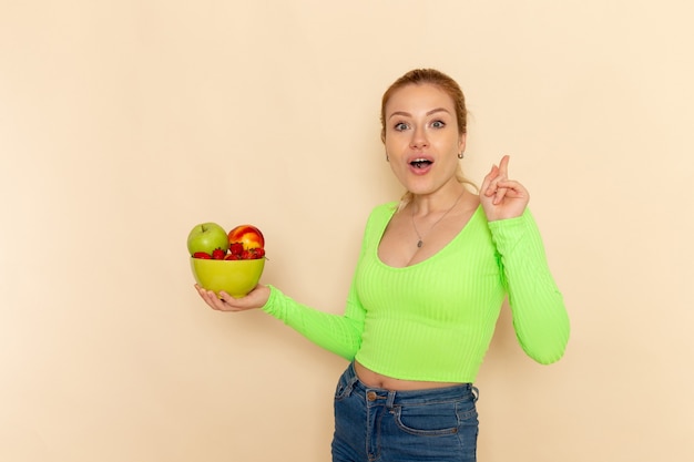 Vista frontal joven hermosa mujer en camisa verde con plato lleno de frutas en la pared de crema ligera modelo de fruta pose de mujer