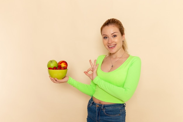 Foto gratuita vista frontal joven hermosa mujer en camisa verde con plato lleno de frutas en la pared de crema ligera modelo de fruta pose de mujer