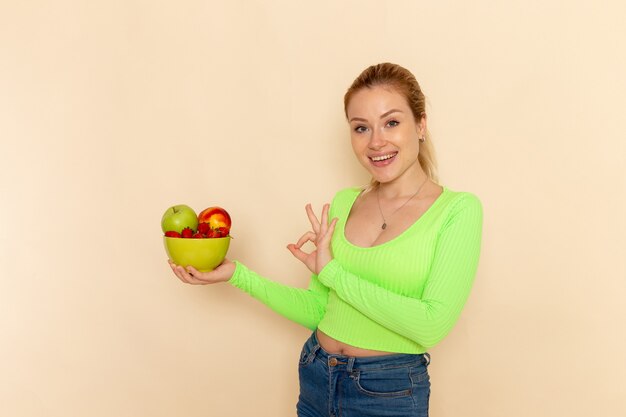 Vista frontal joven hermosa mujer en camisa verde con plato lleno de frutas en la pared de crema ligera modelo de fruta pose de mujer