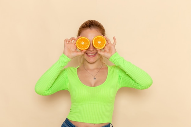 Vista frontal joven hermosa mujer en camisa verde que cubre sus ojos con naranjas en la pared crema modelo de fruta mujer suave