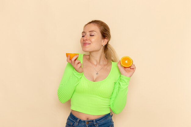 Vista frontal joven hermosa mujer en camisa verde con rodajas de naranja que huelen en la pared de crema modelo de fruta mujer suave