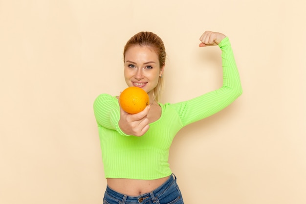 Vista frontal joven hermosa mujer en camisa verde sosteniendo naranja fresca flexionando sobre pared crema modelo de fruta mujer suave