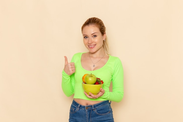 Vista frontal joven hermosa mujer en camisa verde sosteniendo plato con frutas sonriendo en la pared crema ligera modelo de fruta mujer pose dama