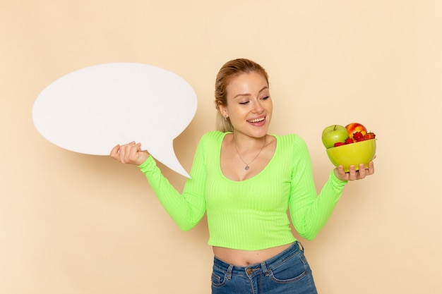 Foto gratuita vista frontal joven hermosa mujer en camisa verde sosteniendo un plato lleno de frutas con un gran cartel blanco en la pared crema mujer modelo de fruta