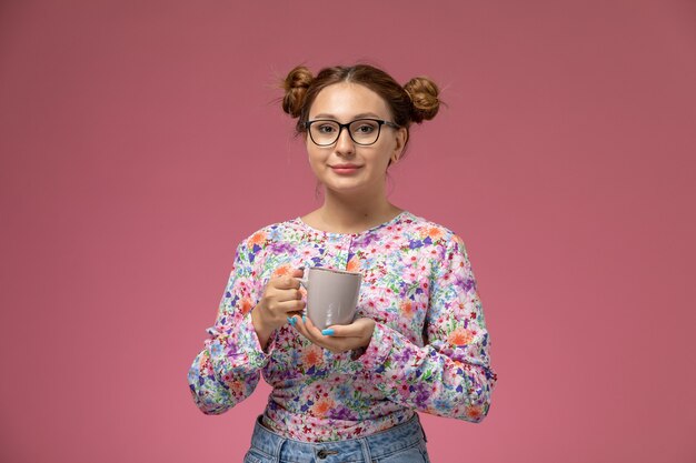 Vista frontal joven hermosa mujer en flor diseñada camisa y jeans sosteniendo una taza de té con una sonrisa sobre fondo rosa