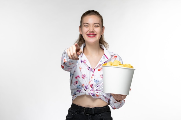 Vista frontal joven hermosa mujer sosteniendo la cesta con patatas cips viendo la película con risas en la superficie blanca