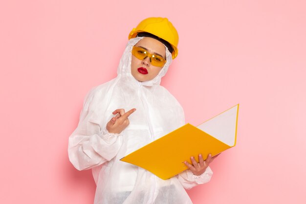 Vista frontal joven hermosa mujer en traje blanco especial con casco protector sosteniendo un archivo amarillo en el traje espacial rosa mujer química