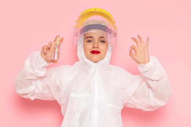 Vista frontal joven hermosa mujer en traje blanco especial con casco protector sosteniendo spray con sonrisa en el espacio rosa traje especial niña mujer