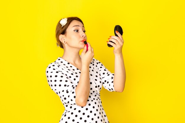 Una vista frontal joven hermosa mujer en vestido de lunares blanco y negro haciendo maquillaje sobre fondo amarillo ropa moda rimel cepillo lápiz labial