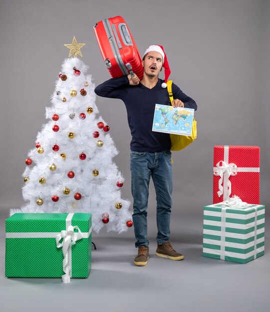 Vista frontal joven hombre de Navidad con mochila amarilla sosteniendo un mapa y una maleta cerca del árbol de Navidad en gris aislado