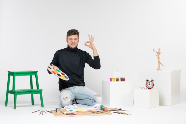 Vista frontal joven hombre sosteniendo pinturas y borla para dibujar en pinturas de pared blanca pintura en color artista arte dibujar imagen
