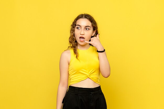 Vista frontal de la joven imitando la llamada telefónica en la pared amarilla