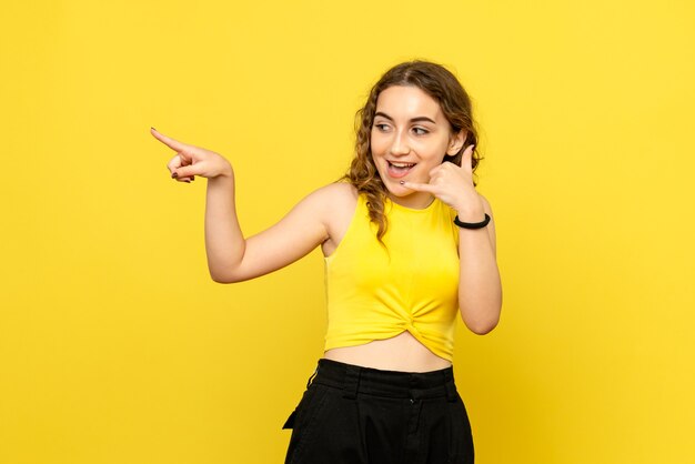 Vista frontal de la joven imitando la llamada telefónica en la pared amarilla